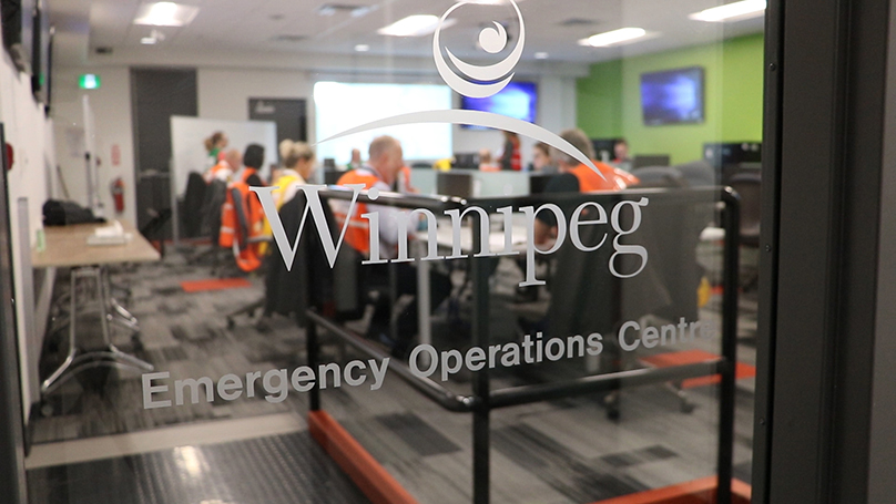 The blurred view through a window at a room full of people working on computers. A decal on the window shows the City of Winnipeg logo and Emergency Operations Centre