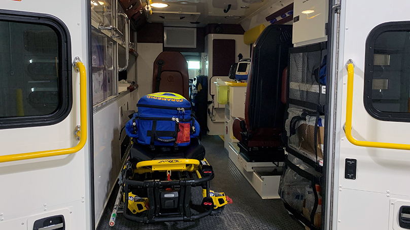 The inside of an ambulance from the back, the doors are open and inside the ambulance is a stretcher with a blue medical bag sitting on top