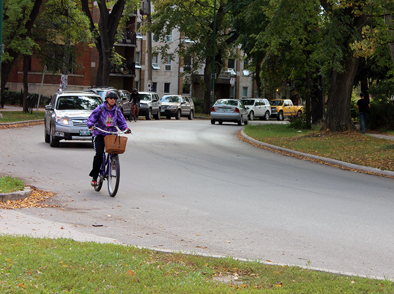 person biking on road with buildings on either side and vehicle behind them