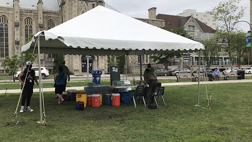 A white tent set up in front of the street on a grassy area with a table set up inside, large water containers sit inside and 5 people are inside the tent