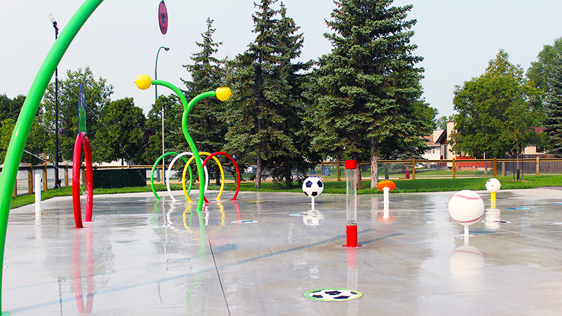 A spray pad with yellow, green, and red fixtures. Soccer ball and baseball fixtures are also shown.