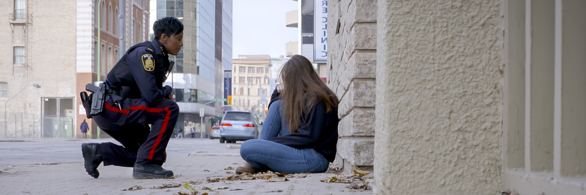 Female police officer kneels in front of a young woman who is sitting on the street