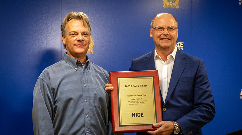 Two men stand together holding a plaque in front of a blue backdrop.