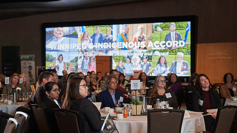 People sit at tables in front of a banner that says "Winnipeg's Indigenous Accord."