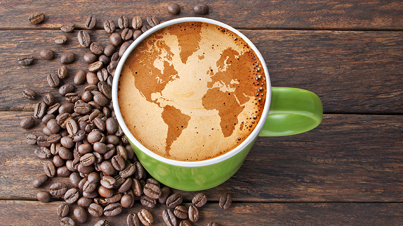 A cup of coffee with a map of the world in it is surrounded by coffee beans on a table