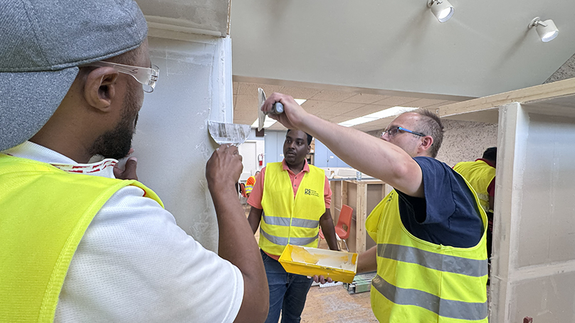 People in safety equipment standing beside a wall watching someone apply putty