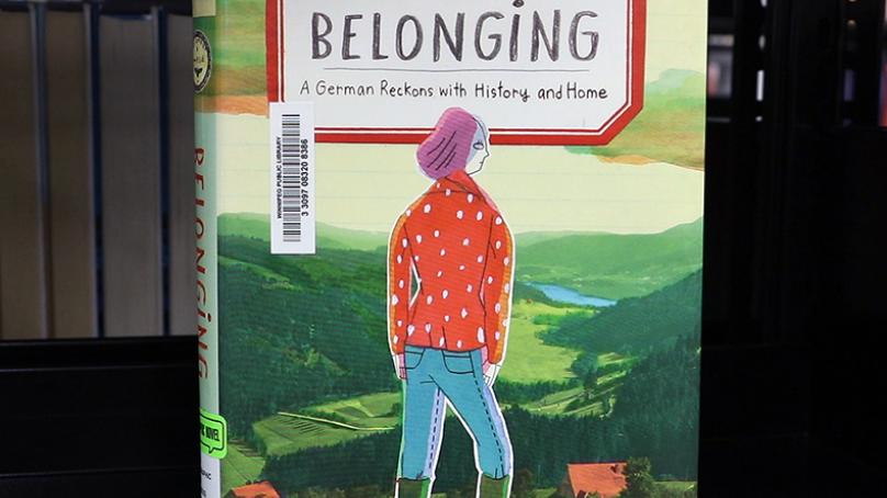  Cover of book Belonging with drawn women with orange jacket and blue jeans and pink beret-type hat