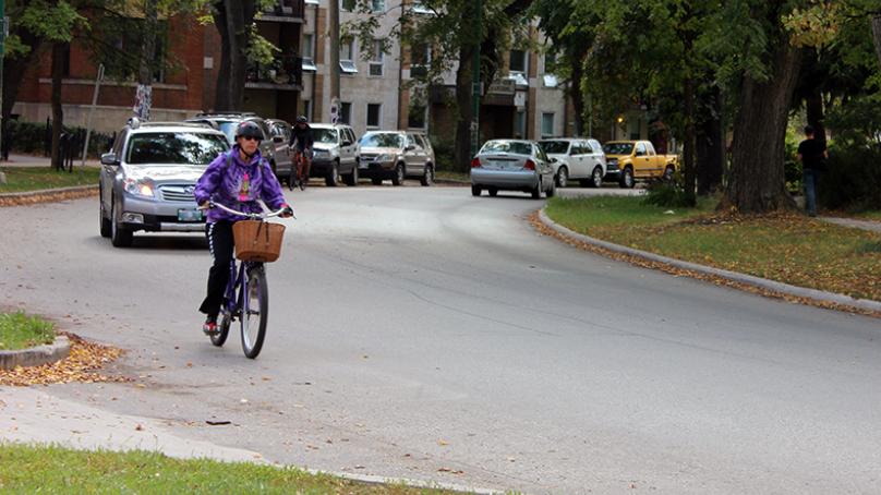 person biking on road with buildings on either side and vehicle behind them