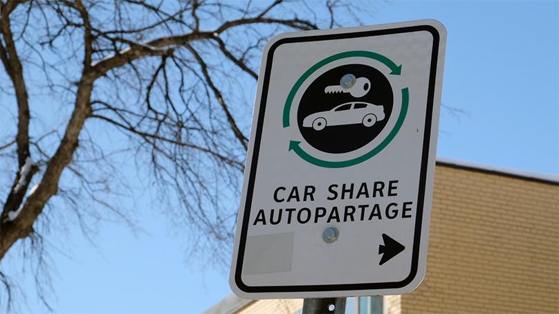 White traffic sign that says Car Share with a black arrow pointing to the right and an icon image of a white car and key on a black circle background with a green circle around it