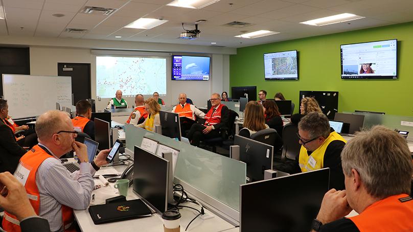 A room filled with people sitting at computers. The people are wearing different coloured vests that are either orange, yellow, red, or green. Unclear images are shown on tv screens on the walls and a map is projected on a smart board at the front of the room.