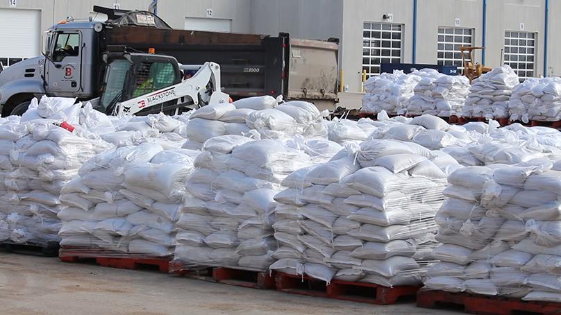 Sandbags are piled on pallets for flood control with skid steer and dump truck behind them