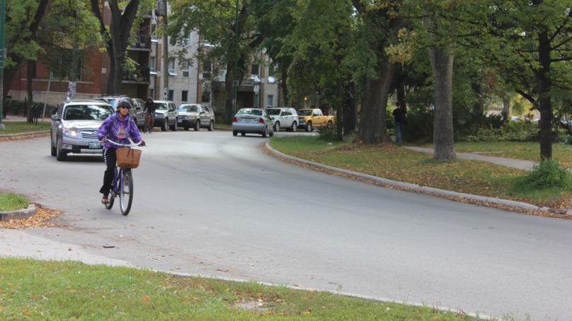 Resident cycling down a street in Wolseley with a car behind them.