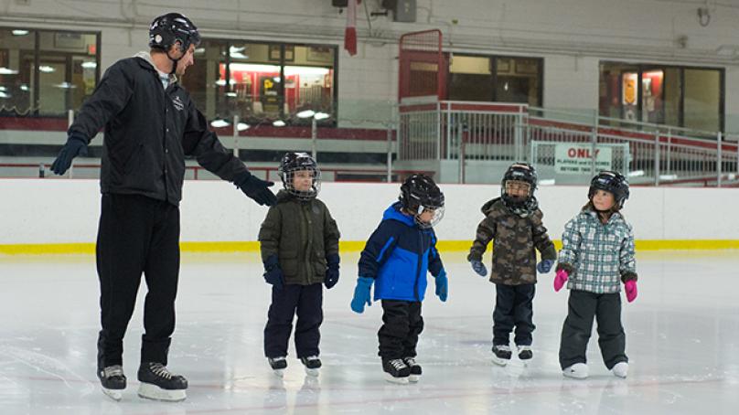 Four kids with helmets and skates standing on a skating rink next to an instructor.