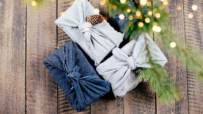 Three small gifts on a wooden plank background. One present is wrapped in blue denim fabric, one is wrapped in grey fabric with a pinecone on top and one is wrapped in grey fabric. An evergreen branch with white lights hangs over the presents.