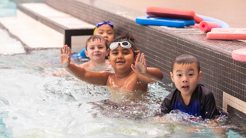 Four children in an indoor pool near the pool edge. Flutter boards and floaties on pool deck