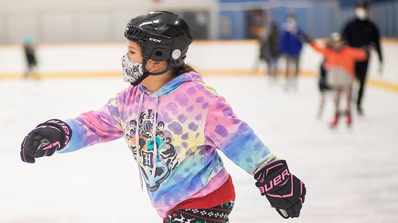 Girl skating in an arena wearing a hockey helmet, gloves. and a tie dye pink and blue sweater. Other kids and adults skating in the background..