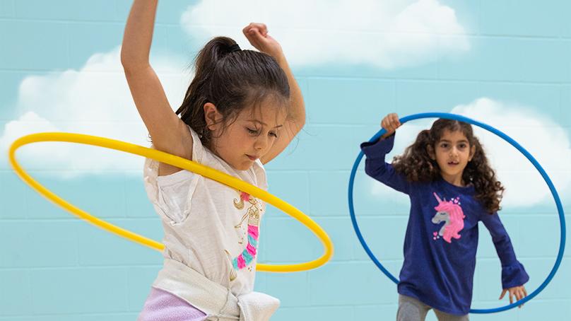 Two girls playing with hula hoops against a painted blue sky with clouds background