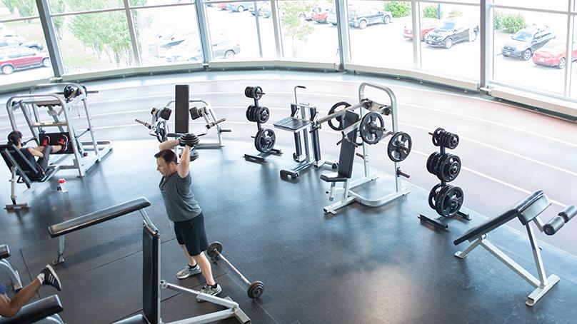 man lifting weights in the weight room and running track area in front of a bank of windows at the Cindy Klassen Recreation Complex