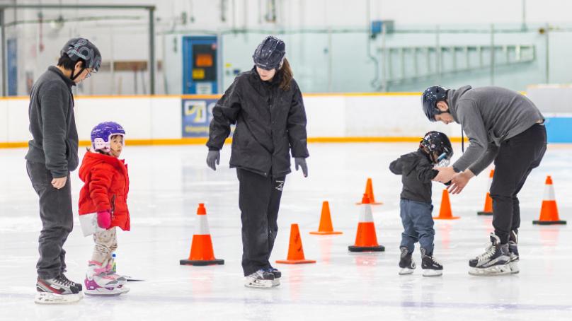 Two dads with their skaters with skate instructor on ice with pylons in line formation at skate lesson