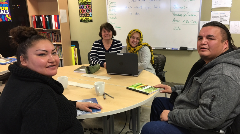 Four individuals sit around a table. Three are students and one is a teacher.
