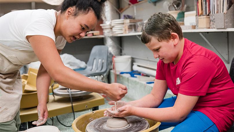 Instructor dropping water onto mound of clay on pottery wheel while student in a red shirt has his hands catching the <a href=