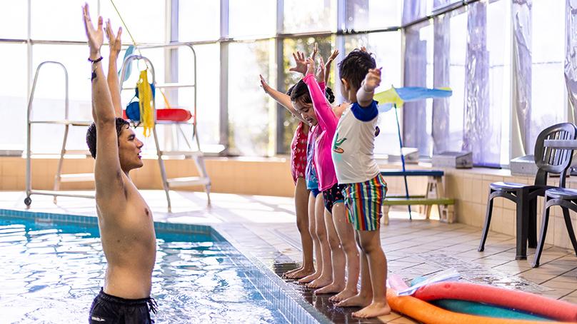 Lineup of preschool swimmers on deck while swim instructor is in water with their arms up over head at swim lesson