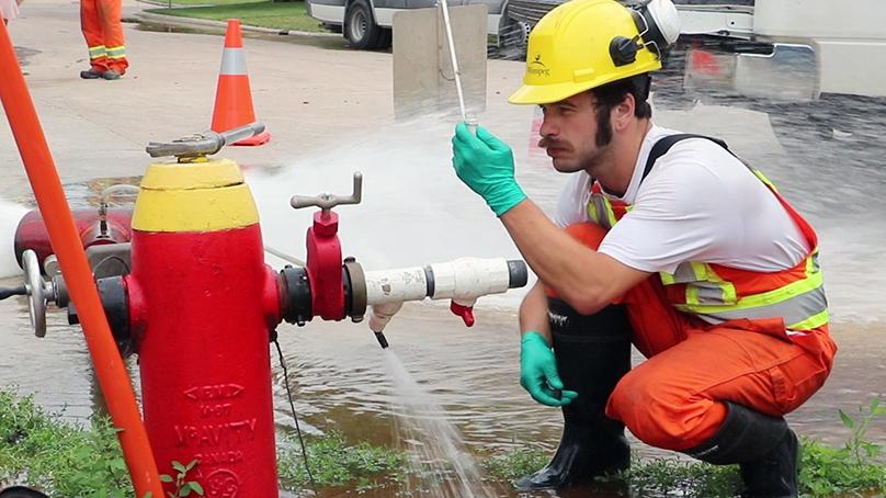 person holding a test tube kneeling beside a fire hydrant spraying water.