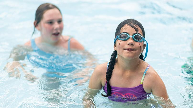 Two girls standing in pool, one has goggles on, during swimming lessons