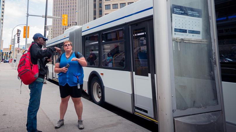 Winnipeg Transit trainer standing at a bus stop helping a passenger.