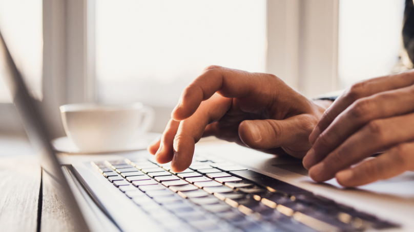 Des mains tapant sur le clavier d'un ordinateur portable. Une tasse de café est posée sur la table à l'arrière-plan.