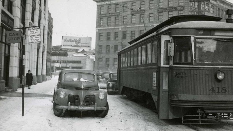 Black and white image of a streetcar beside a parked car in winter 