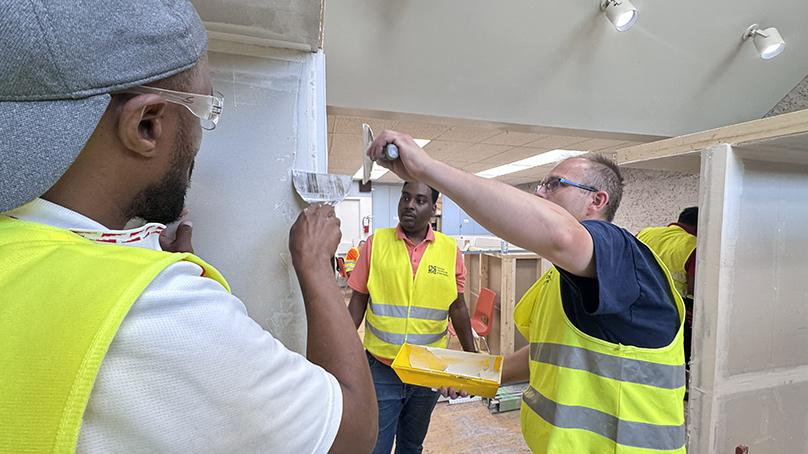 People in safety equipment standing beside a wall watching someone apply putty