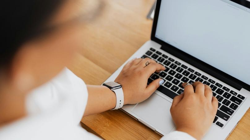 A person sits at a wooden table typing on a laptop.