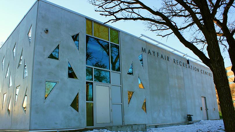 White building with triangle and rectangle windows at dusk during winter. Text on outside says Mayfair Recreation Centre. Framed on the right side by a leafless tree.