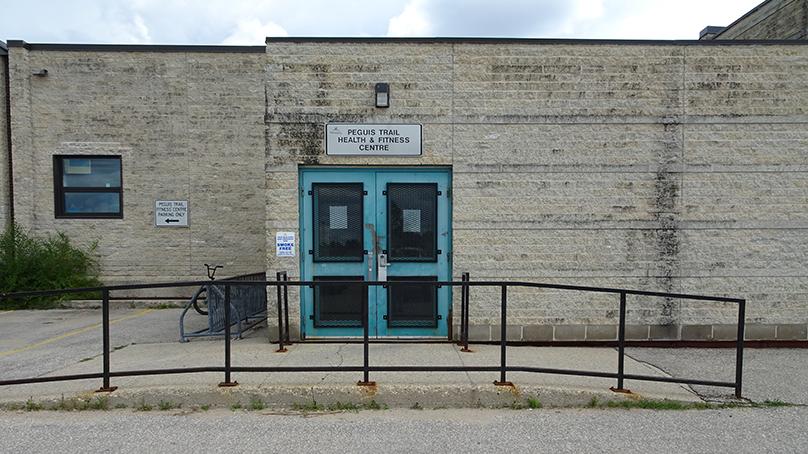 On voit deux portes d’entrée turquoise sous l’enseigne du centre de conditionnement physique Peguis Trail. Une rampe d’accès en pente douce bidirectionnelle est située devant le bâtiment. 