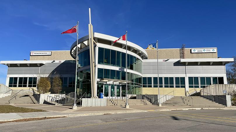 The Manitoban and Canadian flags fly in front of the glass façade of the Pan Am Pool.