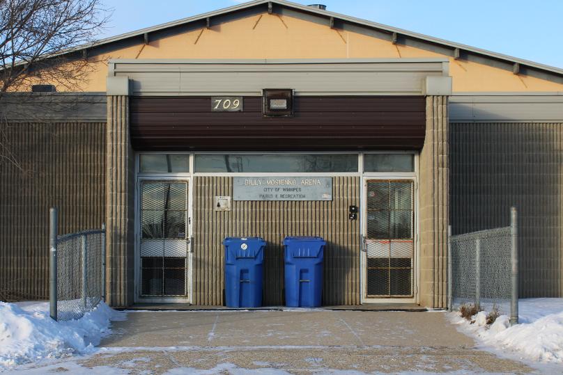 Two blue recycling bins sit next to the front entrance of the Billy Mosienko Arena.